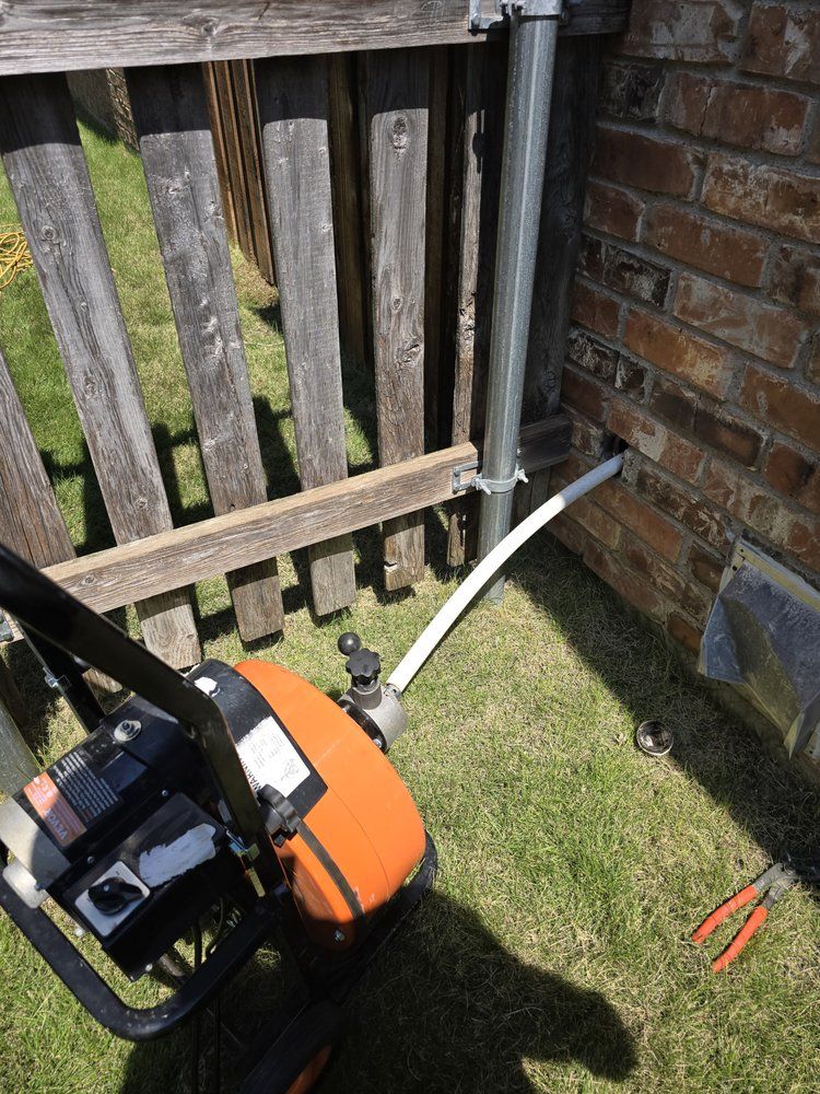 An orange and black pump next to a brick wall and a wooden fence. A white hose extends from the pump into the wall.