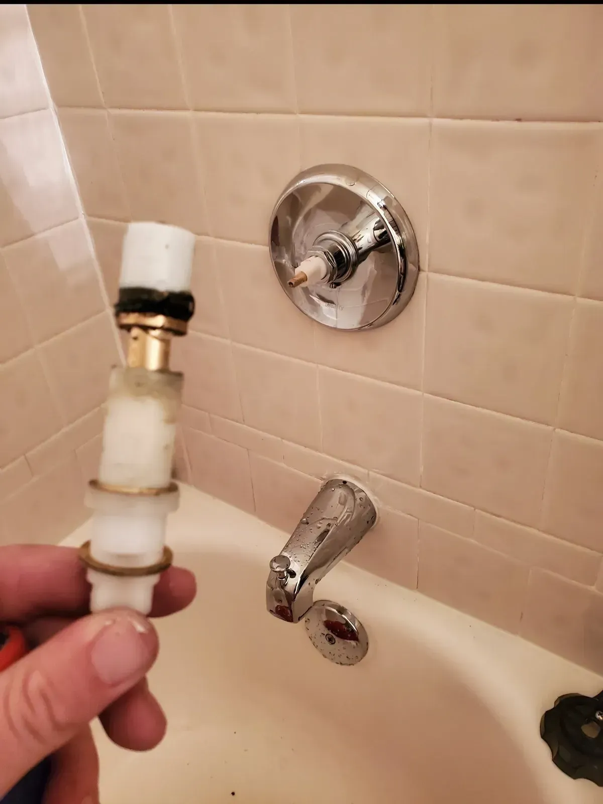 Hand holding a shower faucet cartridge next to a showerhead and faucet. White and chrome, bathroom setting.