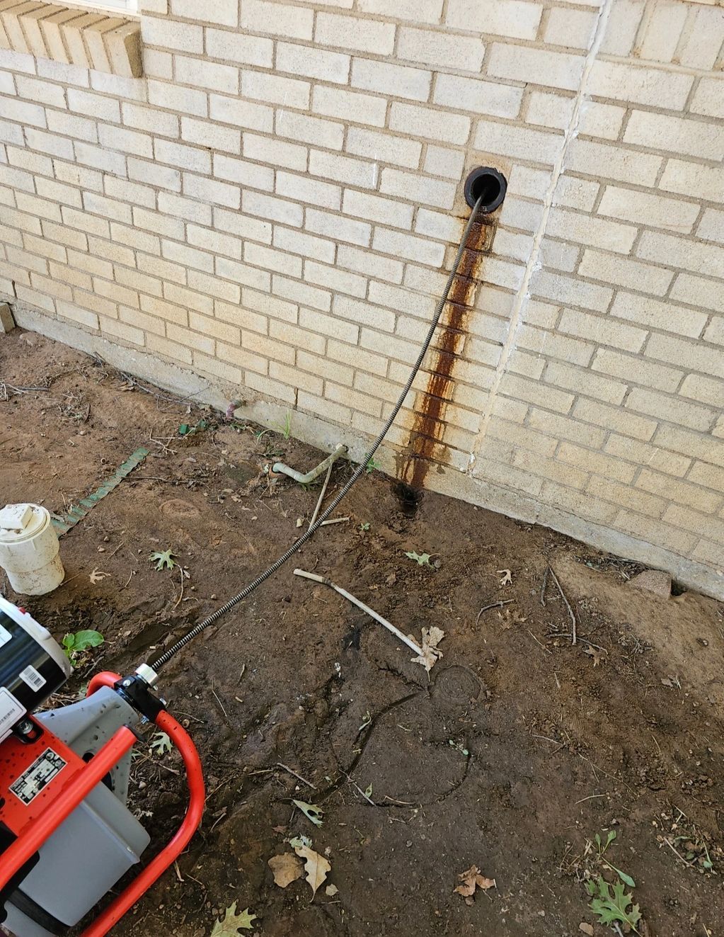 A drain cleaning machine is at a brick wall with a black pipe. Cable extends from the machine into the pipe.