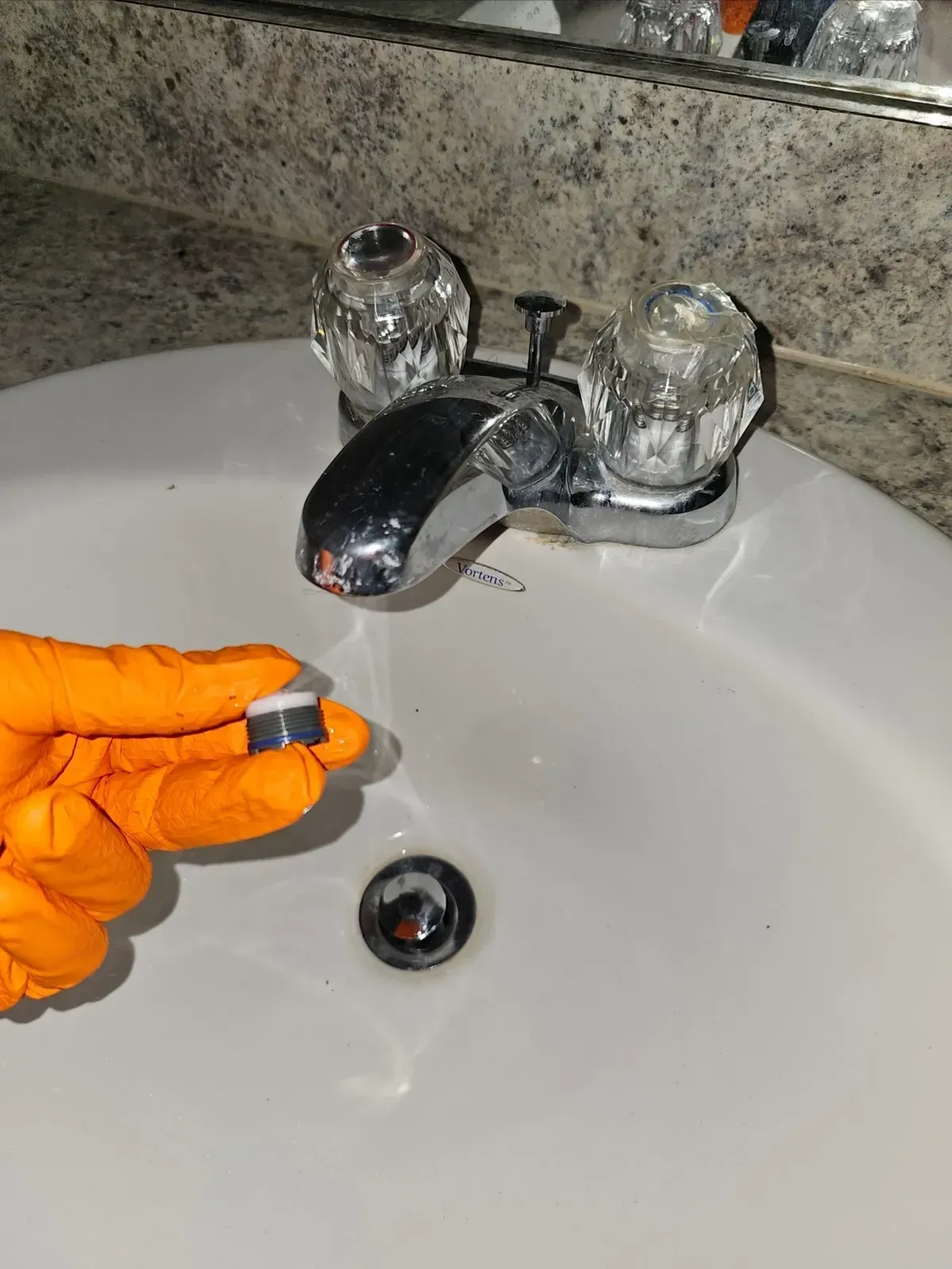 Hand in orange glove holding a faucet aerator near a sink. Chrome faucet with crystal knobs.