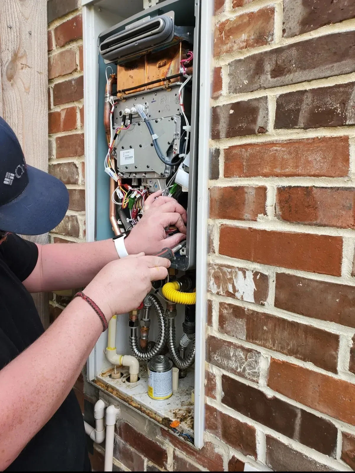 A person repairs a water heater in a brick wall. Wires and pipes are visible.