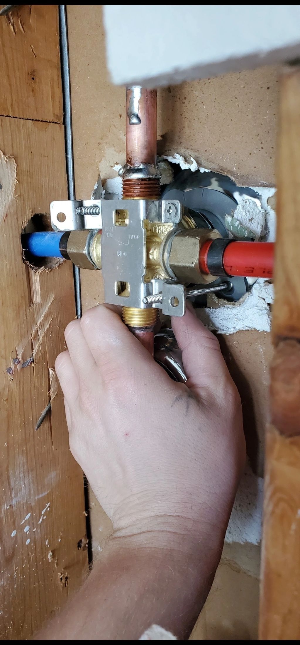 A person's hand adjusting plumbing valves and pipes in a wall. Copper, red, and blue pipes are visible.