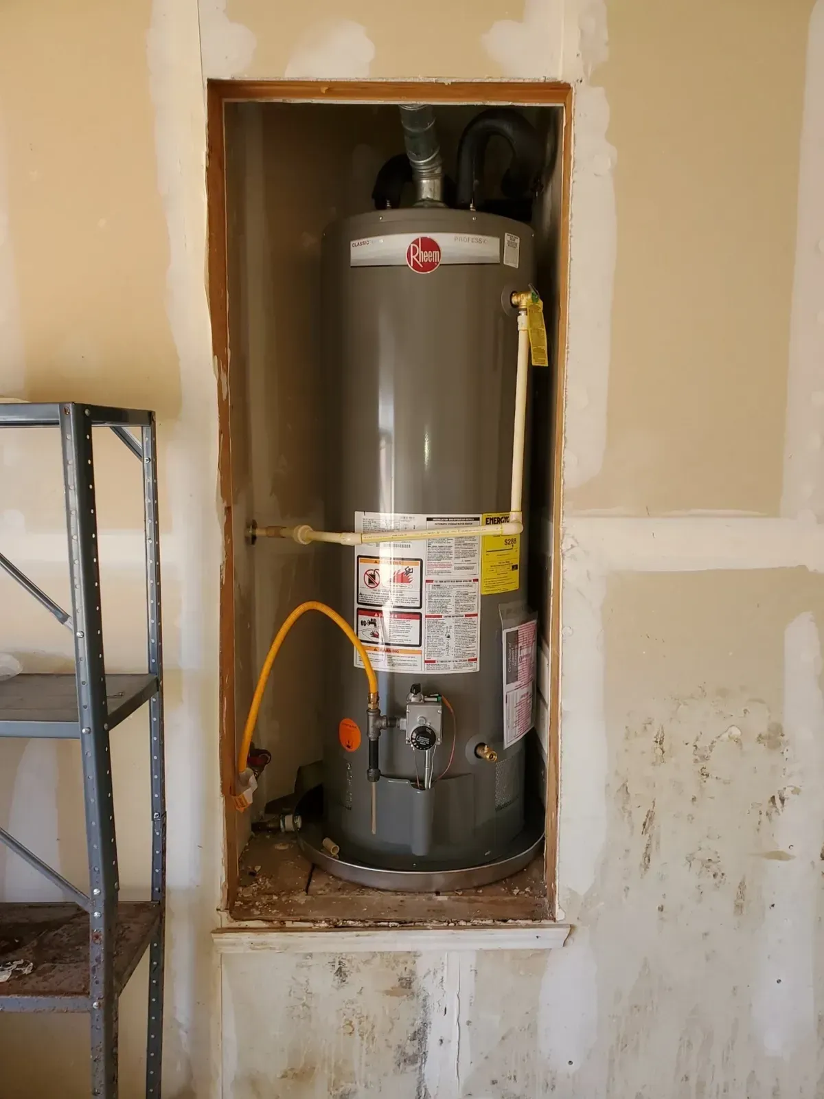 A gas water heater in a wall alcove, surrounded by unfinished drywall and a metal shelf.