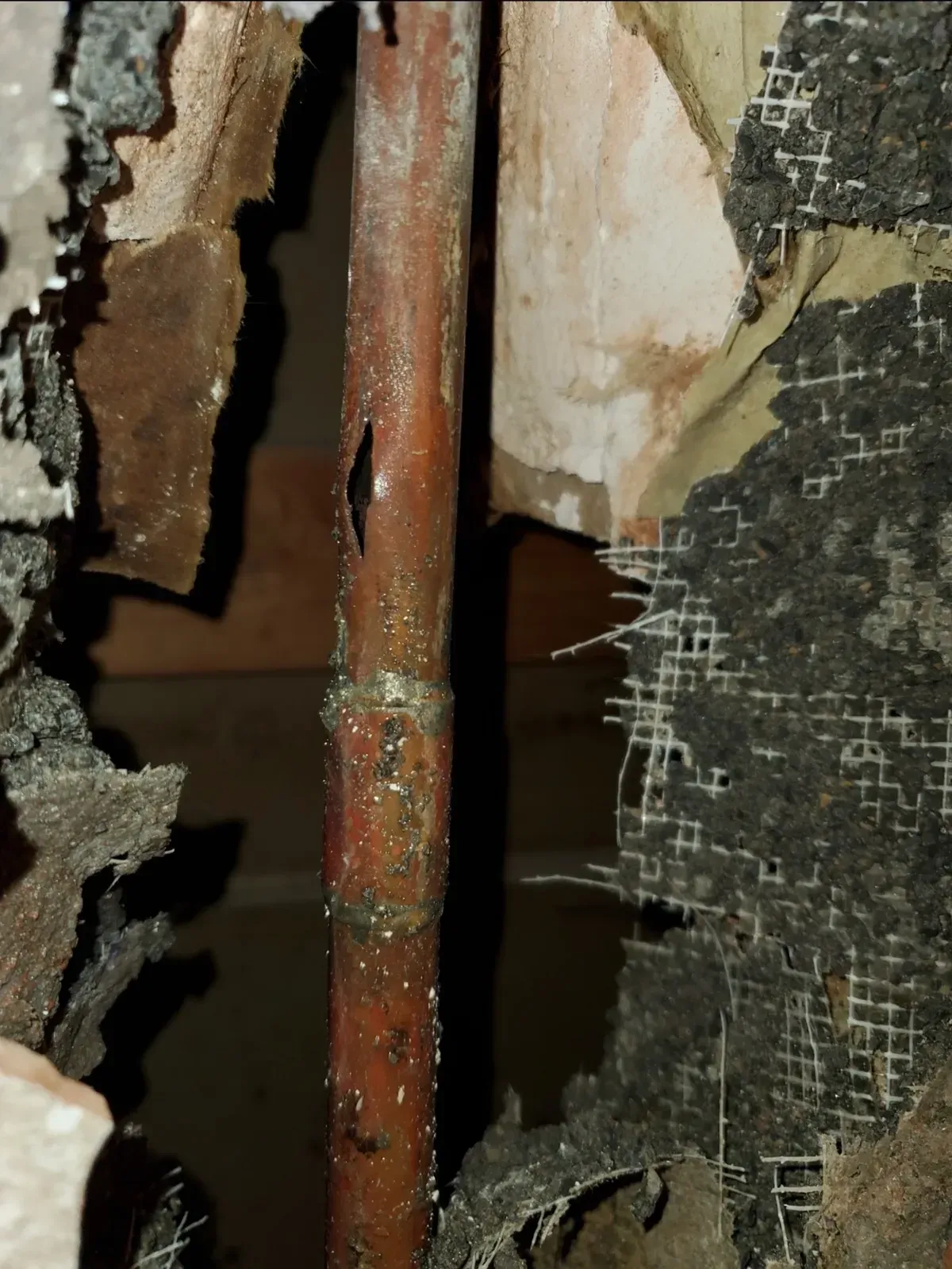 A copper pipe with a hole leaking water, surrounded by damaged wall materials.