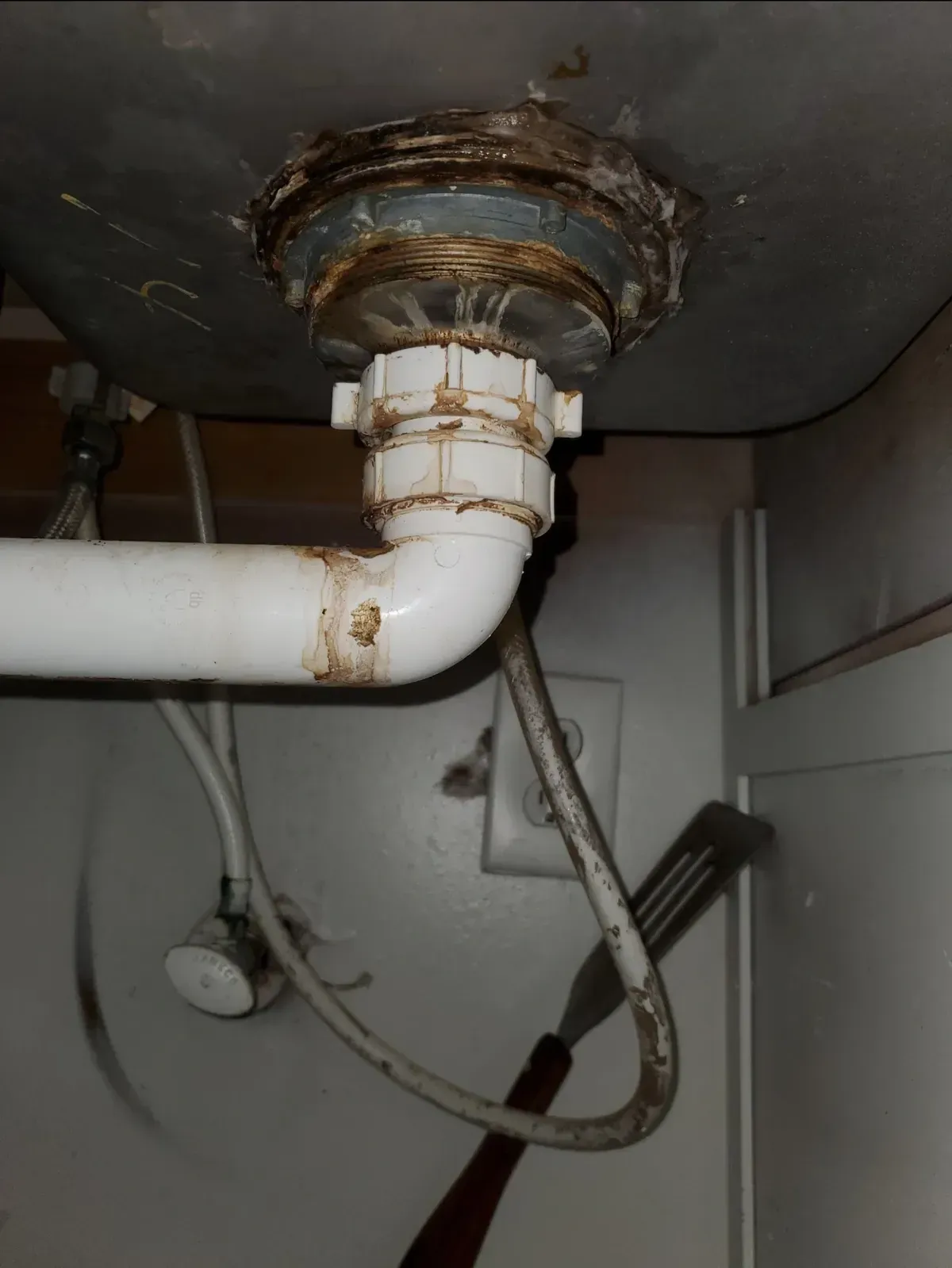 Underside of a kitchen sink with a white drain pipe and fittings. Some rust and debris are visible.
