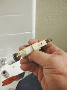 Hand holding a white and brass shower valve stem, likely for repair, against a bathroom setting.
