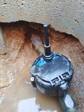 Water meter in a dirt cavity with a black cover, surrounded by mud and a pipe.