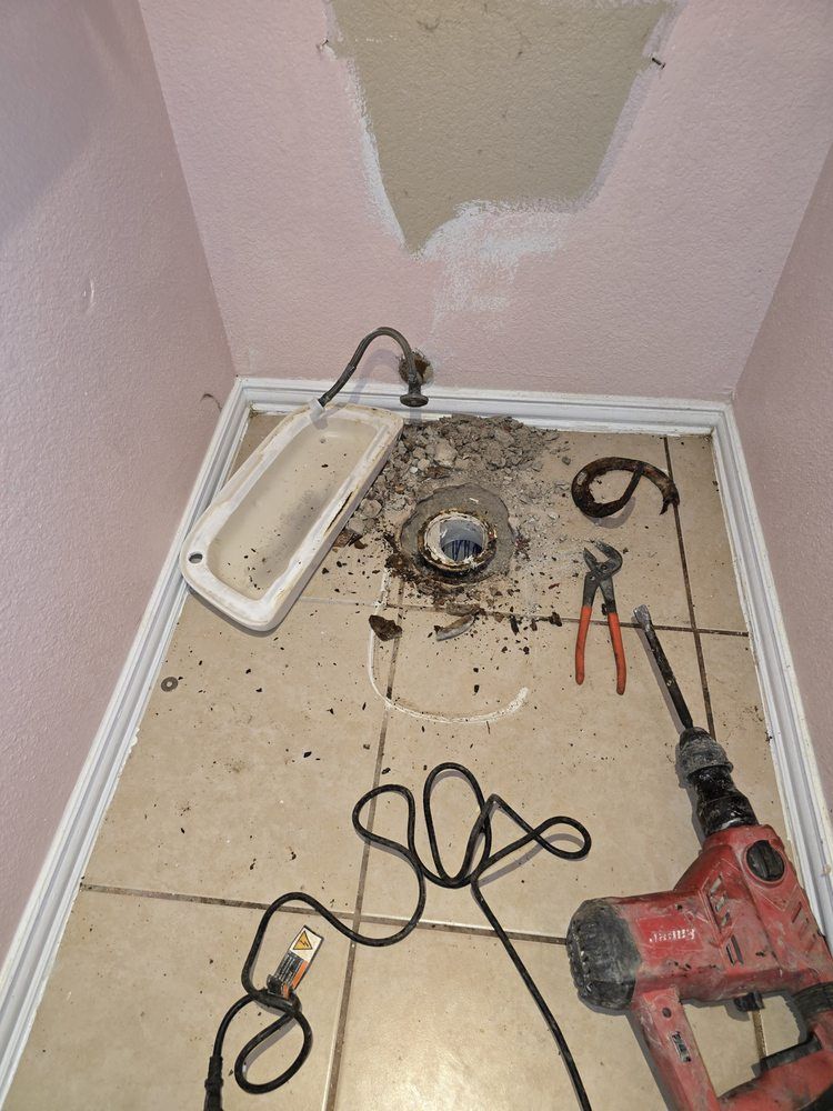 Bathroom floor demolition: broken tiles, plumbing exposed, power tools, and a partly removed ceiling.