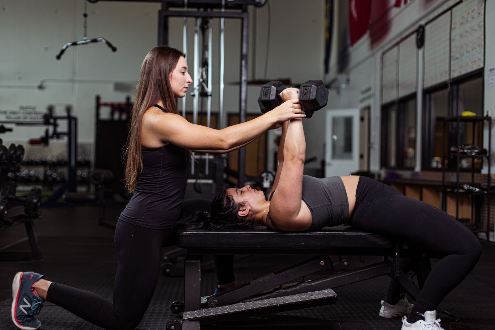 Woman spotter assisting another woman doing dumbbell chest press on a bench in gym.