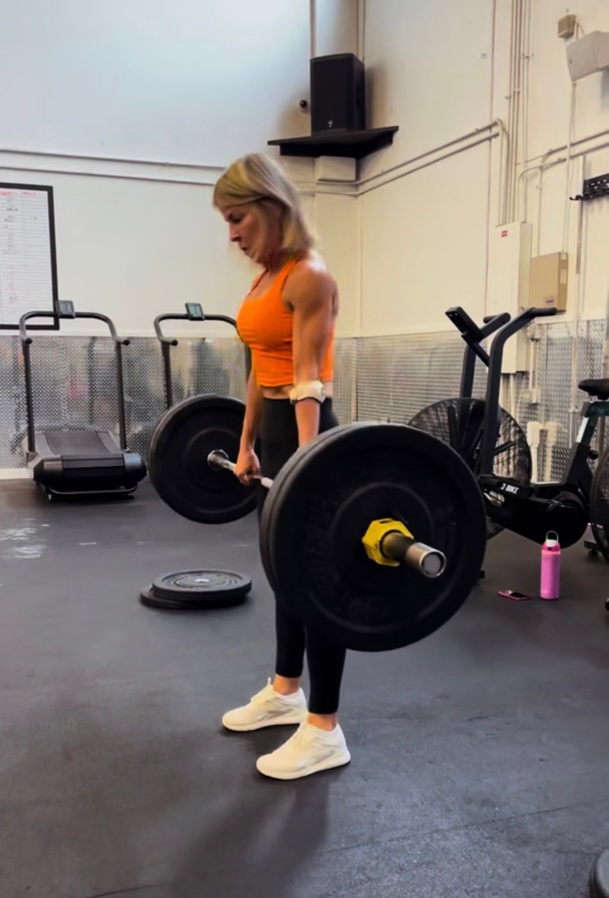 Woman rowing indoors on Concept2 machine. Another person in background. Gym setting.