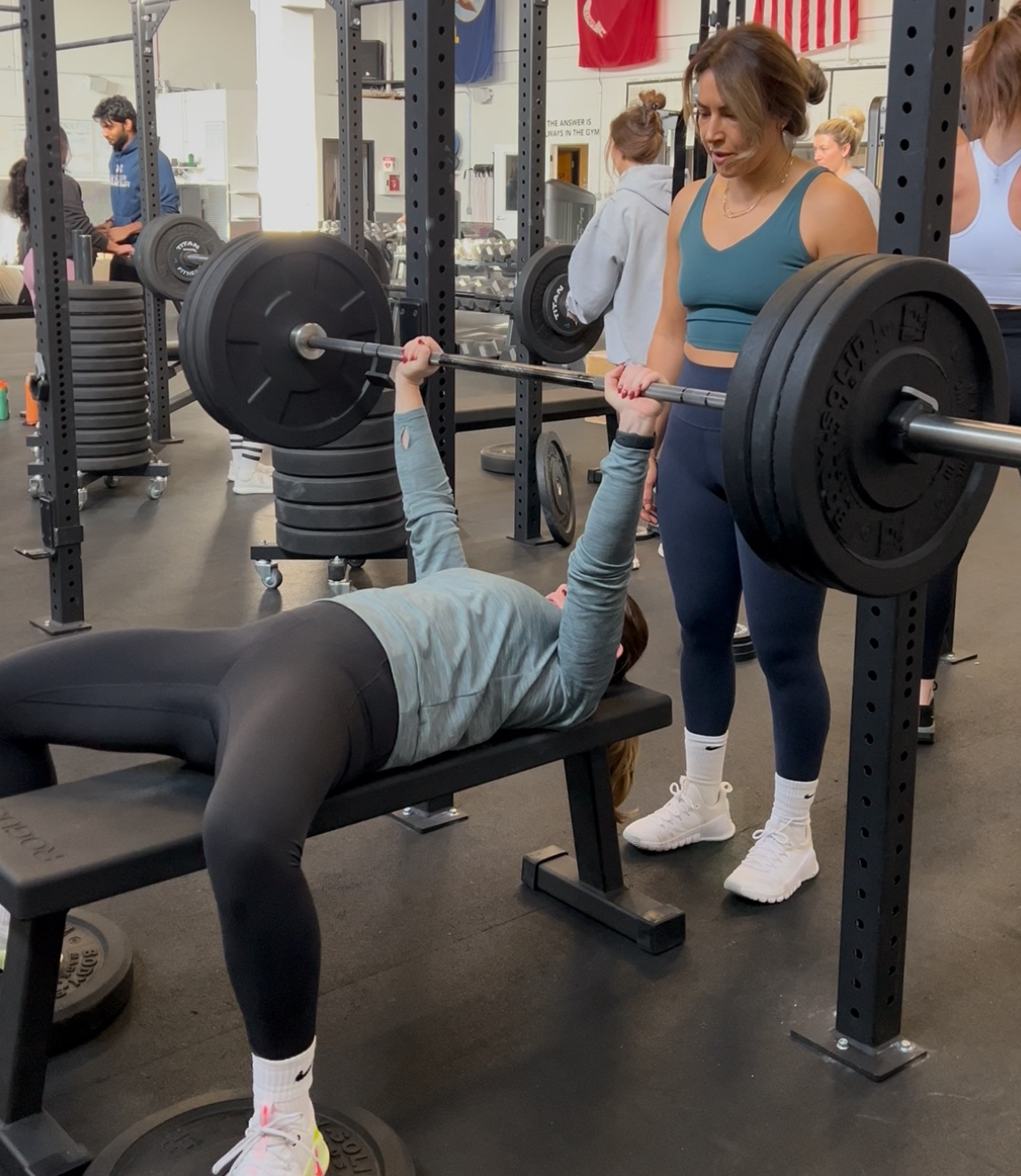 Woman doing kettlebell squat, guided by a trainer in a gym setting.