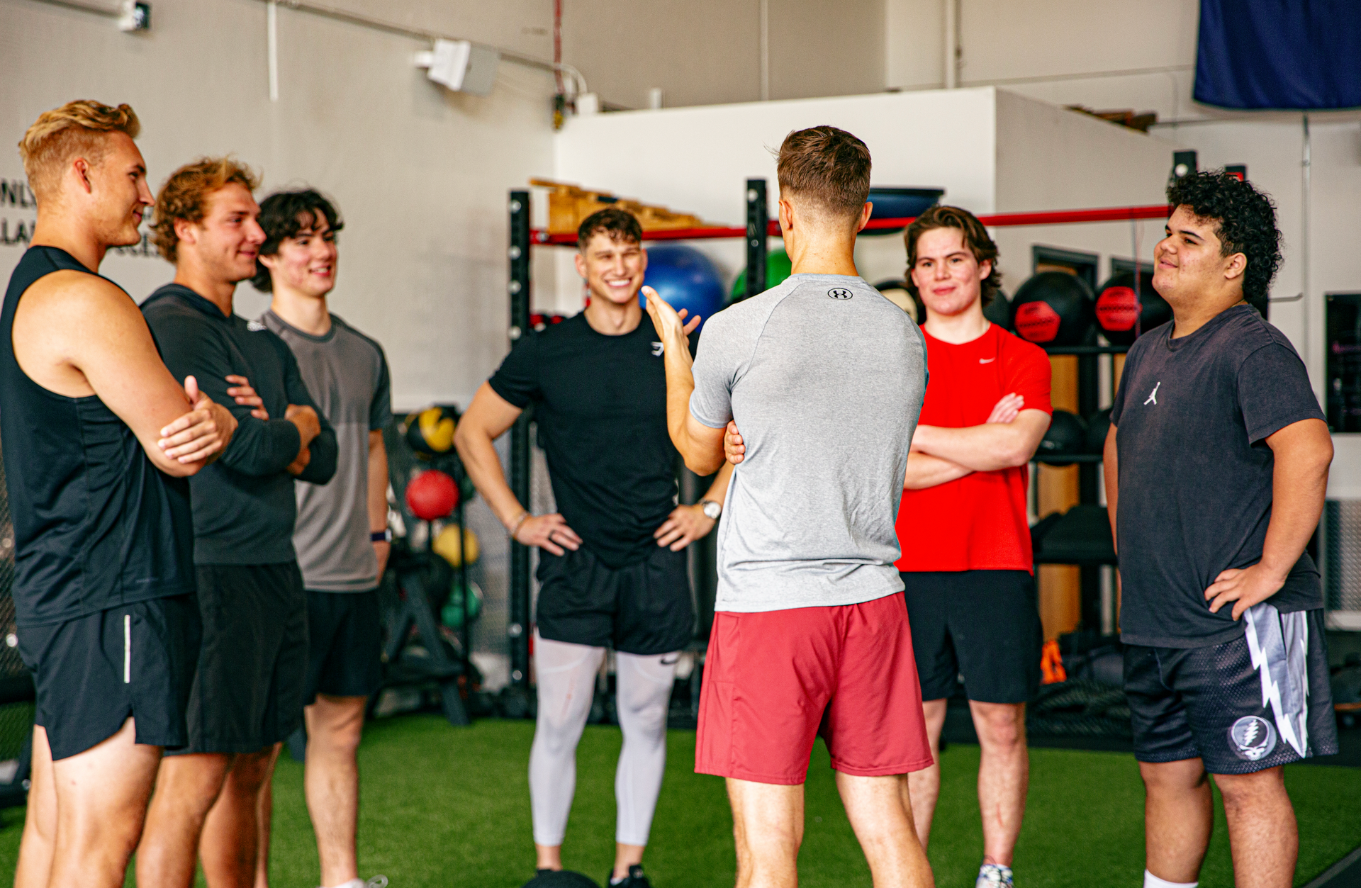 Group of young men in a gym, listening to someone in red shorts.