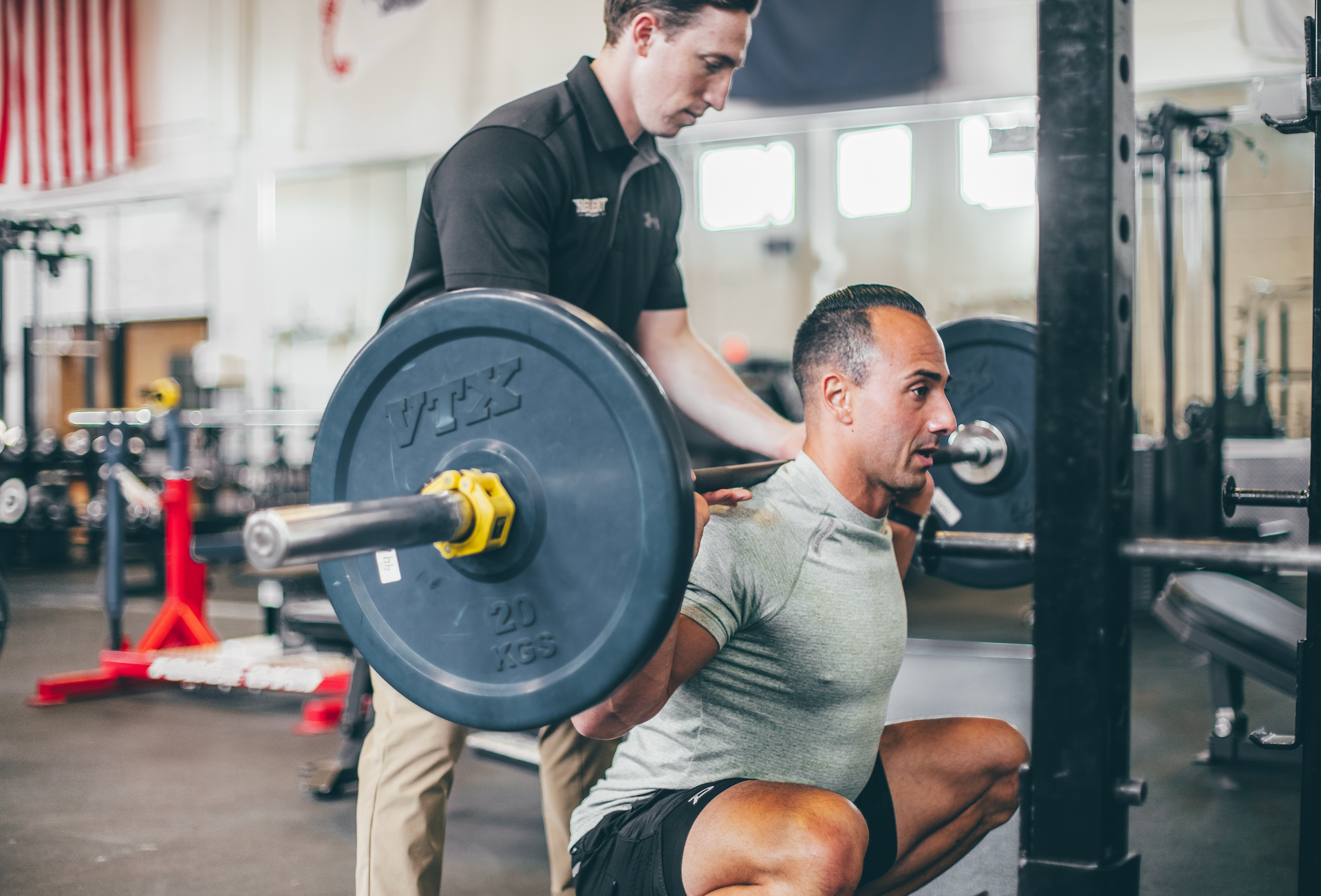 Man doing a squat with a barbell, supervised by a trainer in a gym.