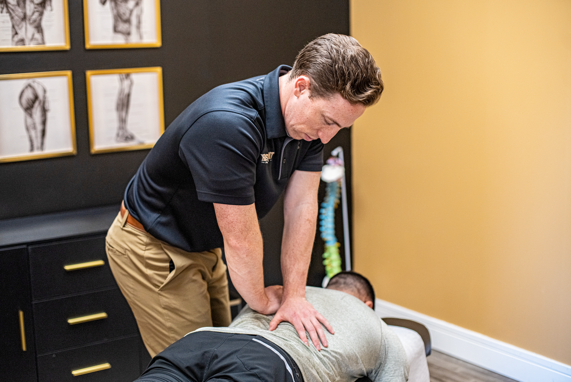 Chiropractor adjusting a patient's back in a clinic. The doctor is in a dark polo shirt, khaki pants.