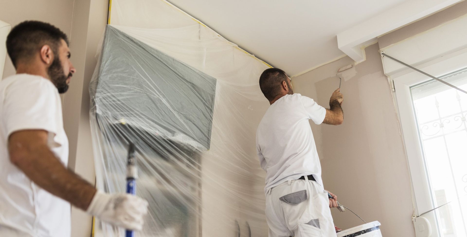 two men prepare and paint a wall in a room