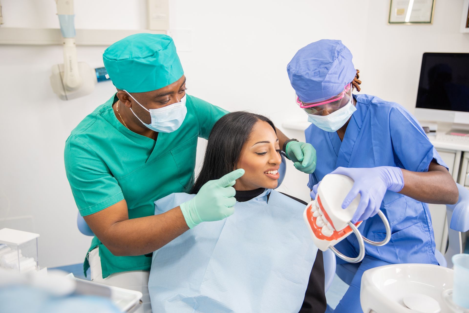 Dentists in scrubs and masks examining a patient in a dental chair, using a model of teeth.