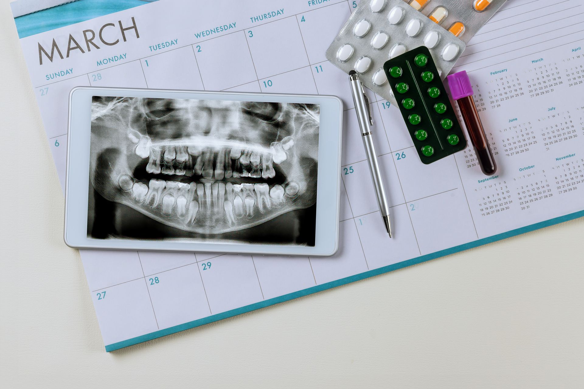 Tablet displaying dental X-ray, surrounded by medication, blood vial, pen, and a March calendar.