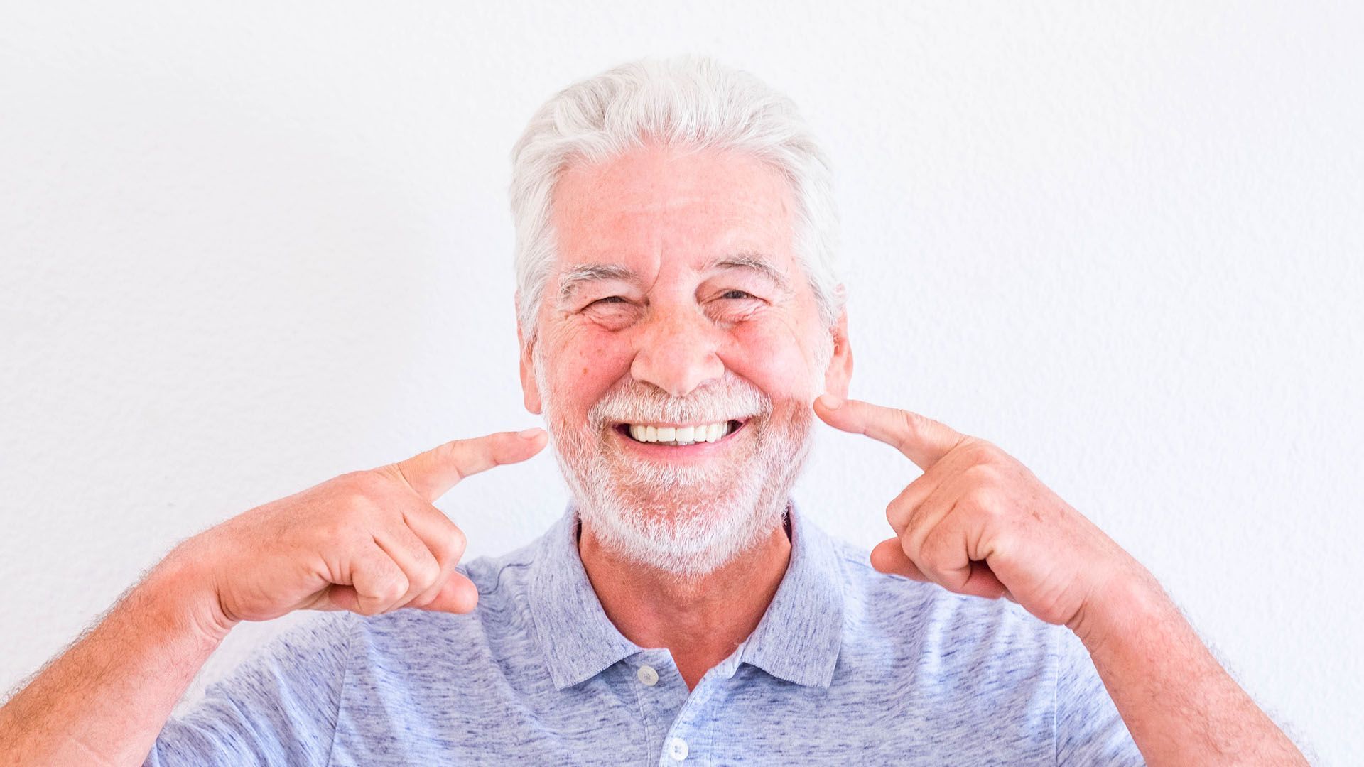 Elderly man with white hair and beard smiling, pointing to his cheeks with both index fingers.