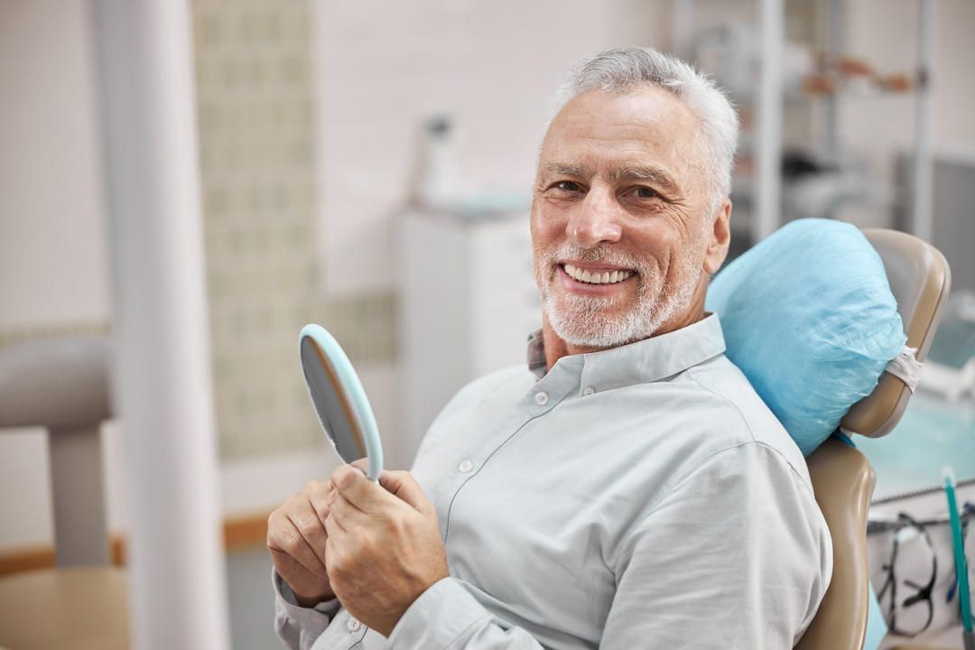 A man is sitting in a dental chair looking at his teeth in a mirror.