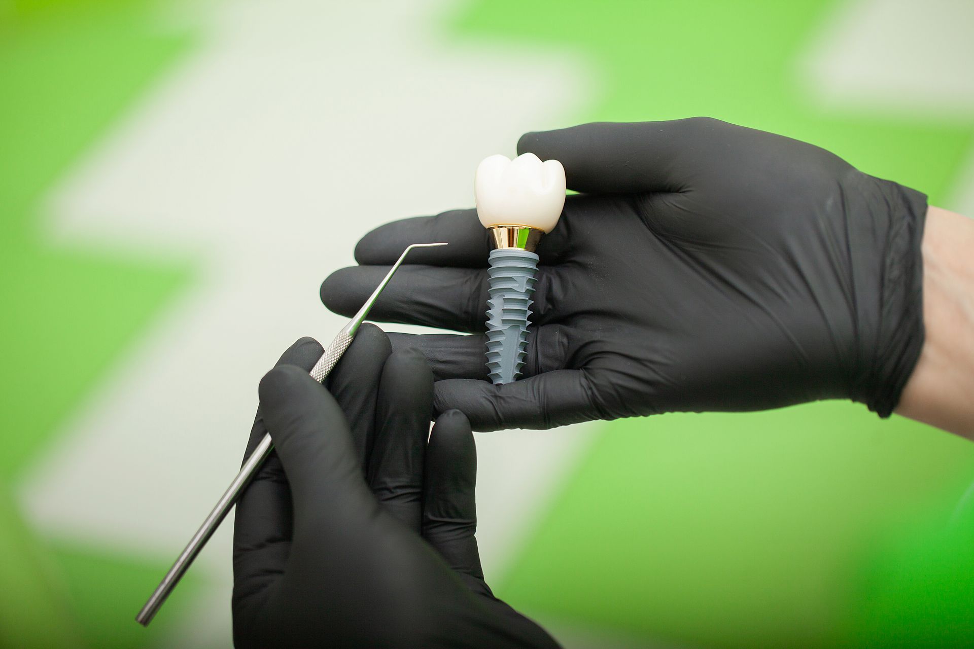 Hands in black gloves hold a dental implant assembly with a white crown and a metallic screw against a green background.