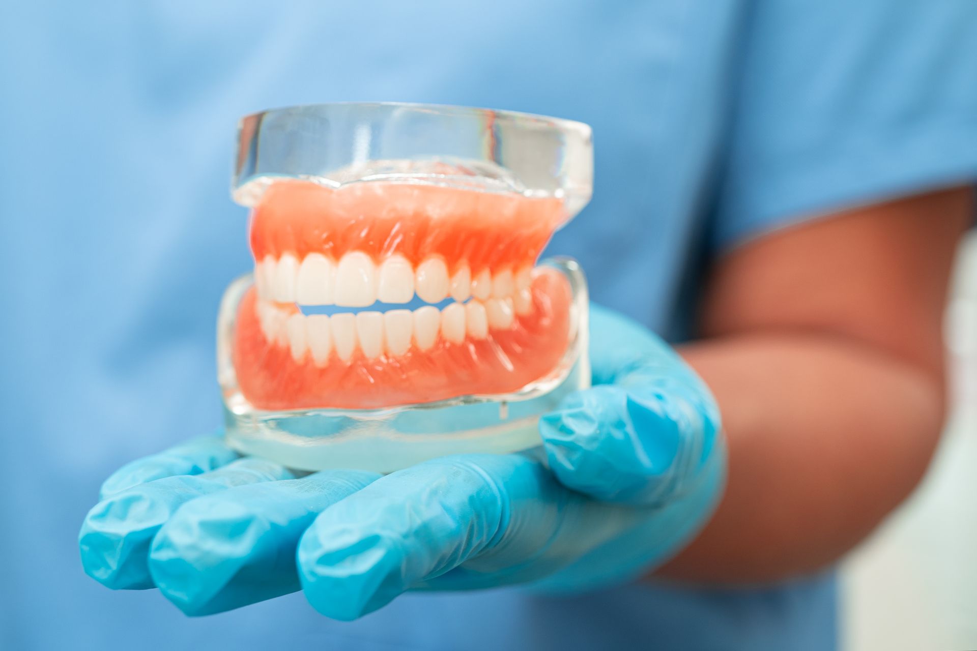 A hand in a blue medical glove holds a set of complete dentures mounted in a clear plastic articulator.