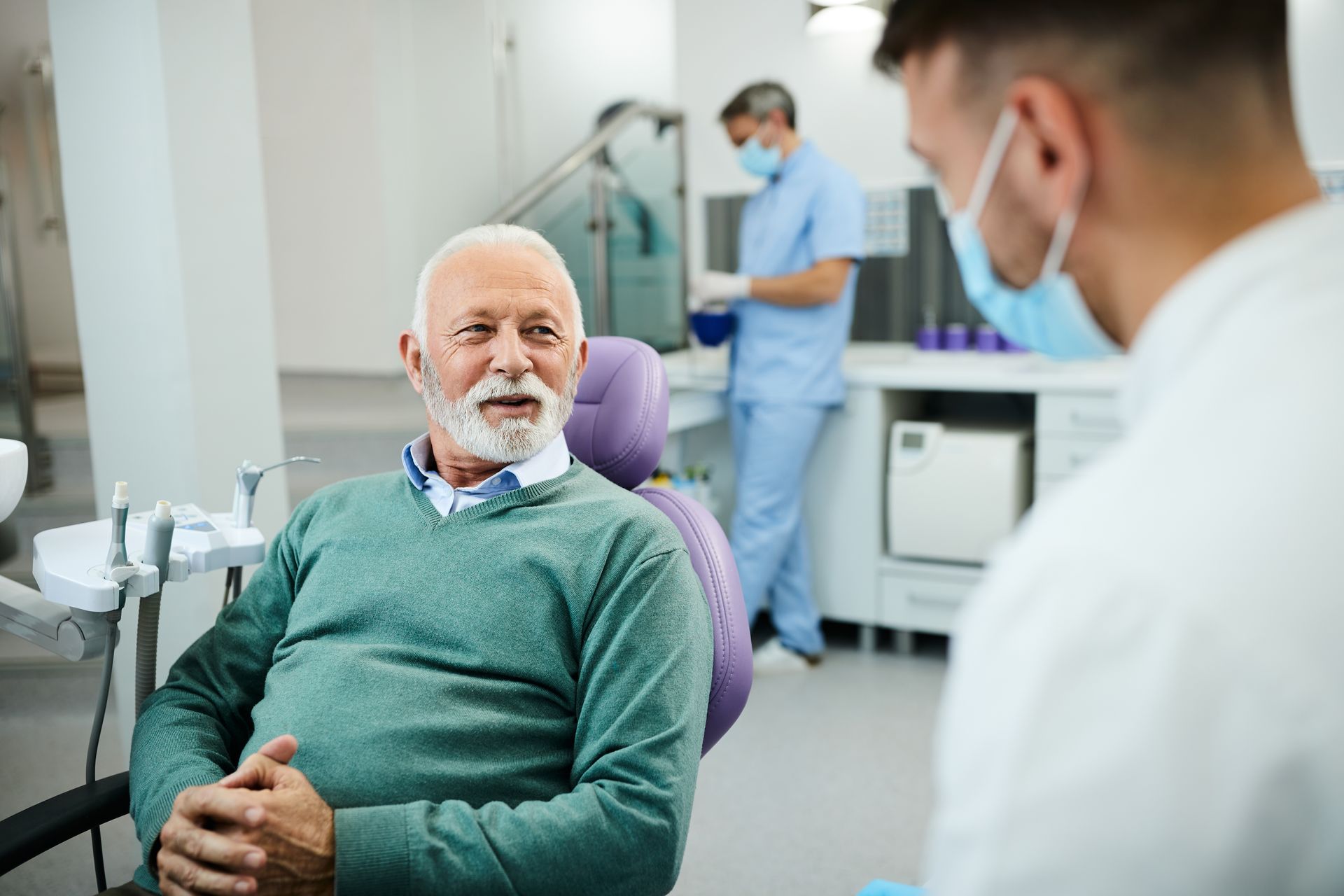 Dentist consulting with a seated patient in a bright clinic, with staff working in the background.
