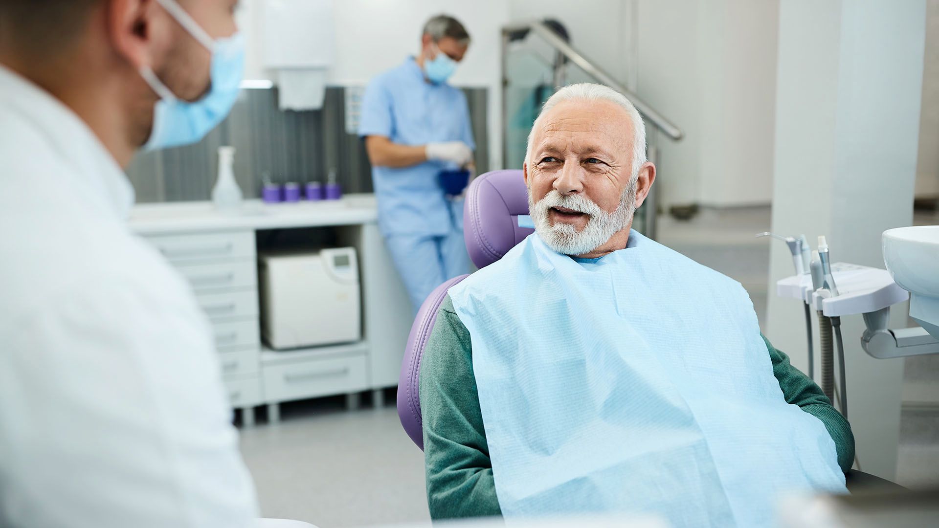 Senior man in dental chair speaking to a masked dentist; dental office setting.