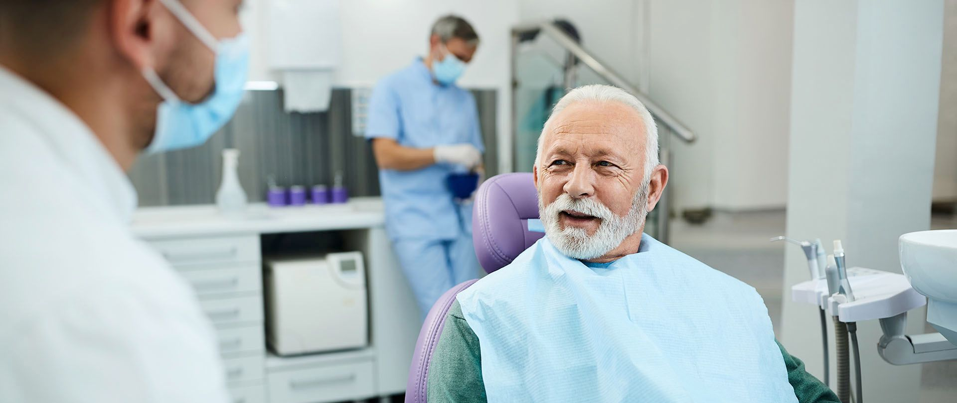 A dentist wearing a mask talks to a seated elderly patient at a clinic.