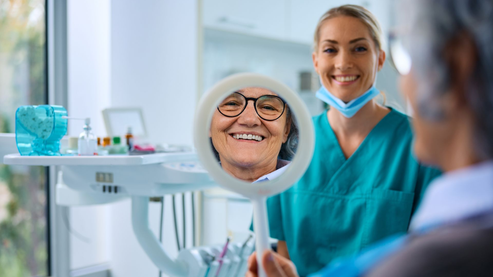 Dentist shows a smiling older patient her teeth in a mirror. Dental office, green scrubs.