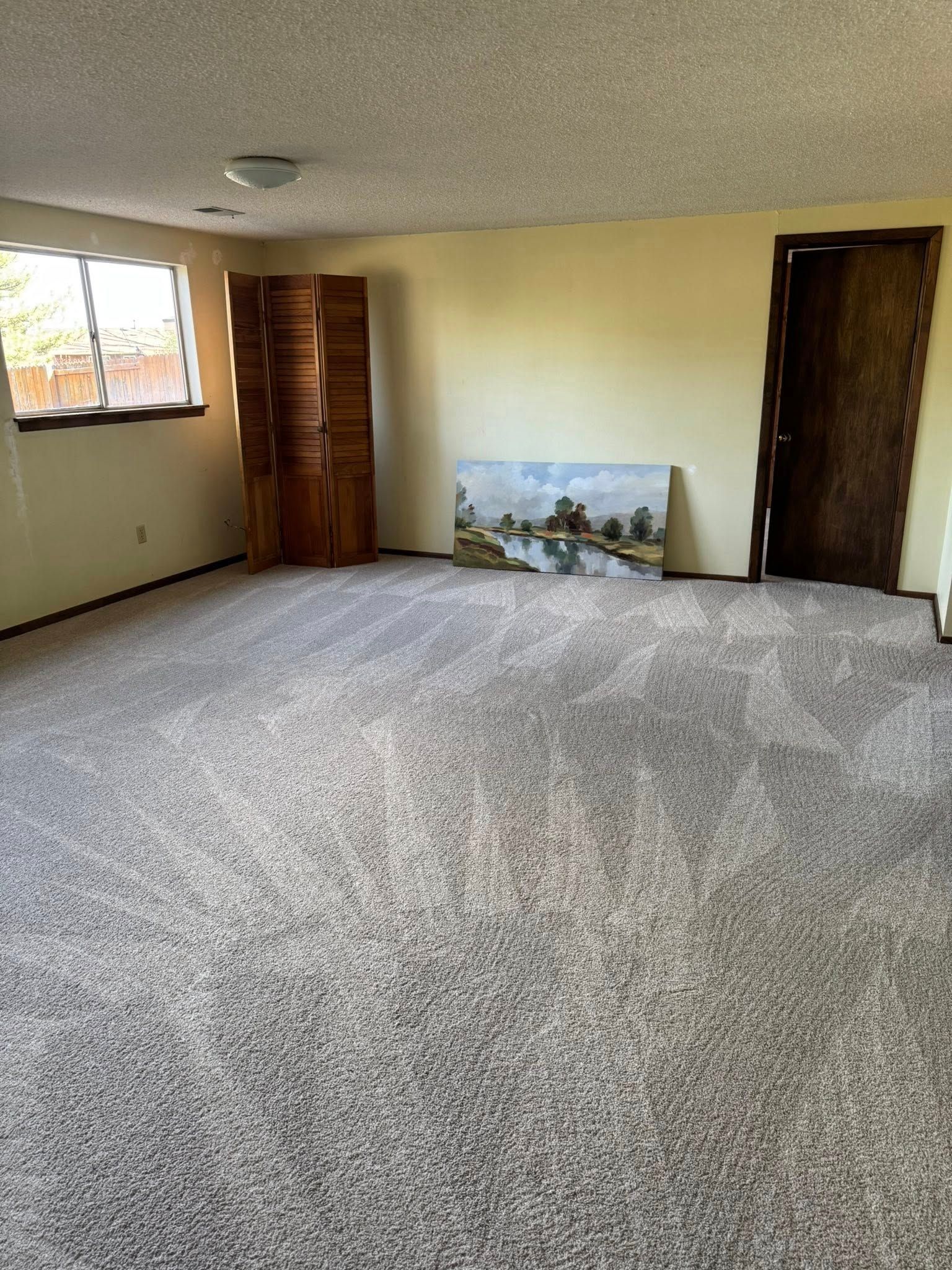 Empty room with beige walls and gray carpet, wooden screen, window, and painting against the wall.