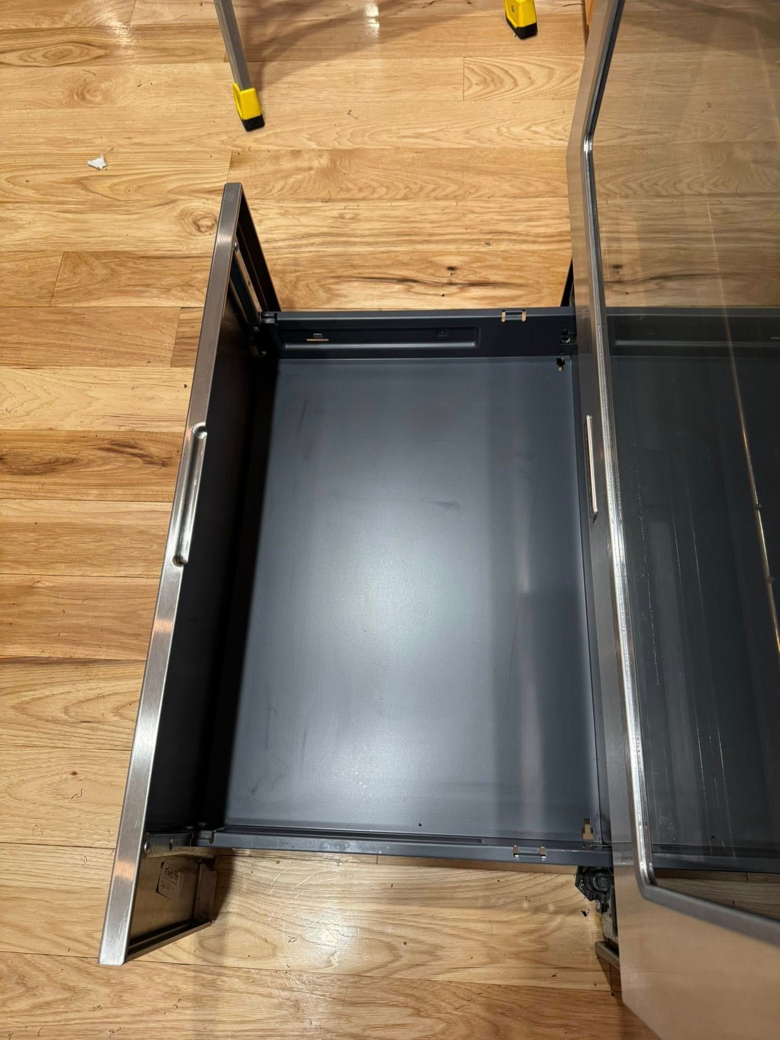 Open, empty, dark gray drawer in a stainless steel cabinet on a wood floor.