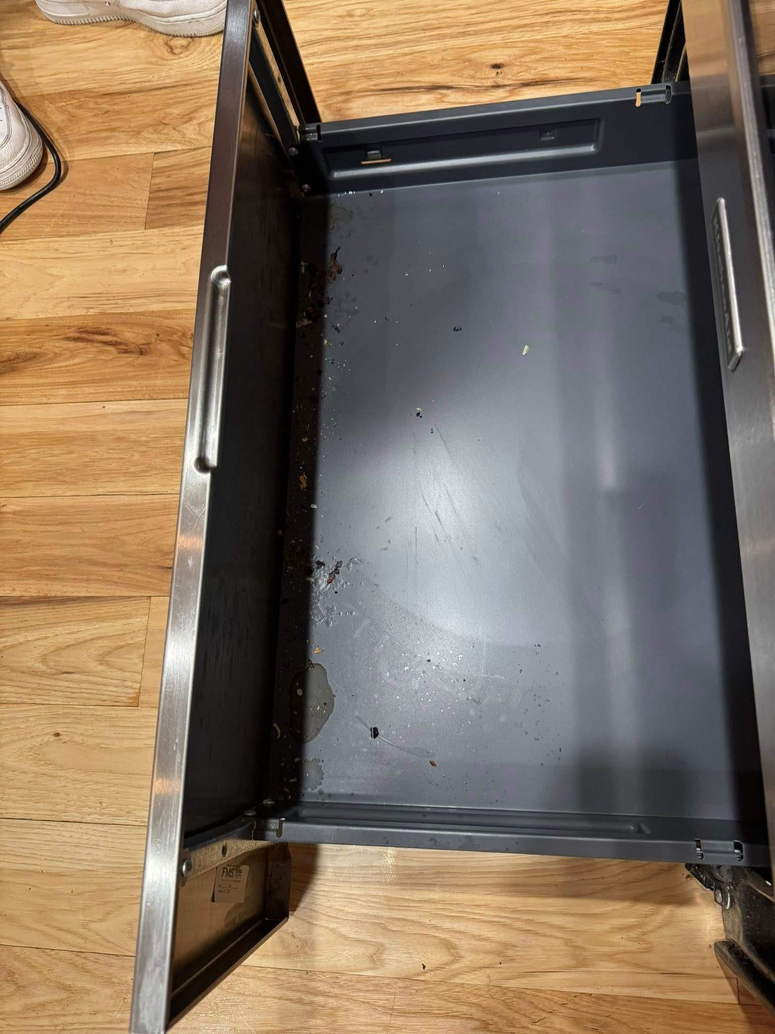 Open metal drawer with dark gray interior, set on a wooden floor.