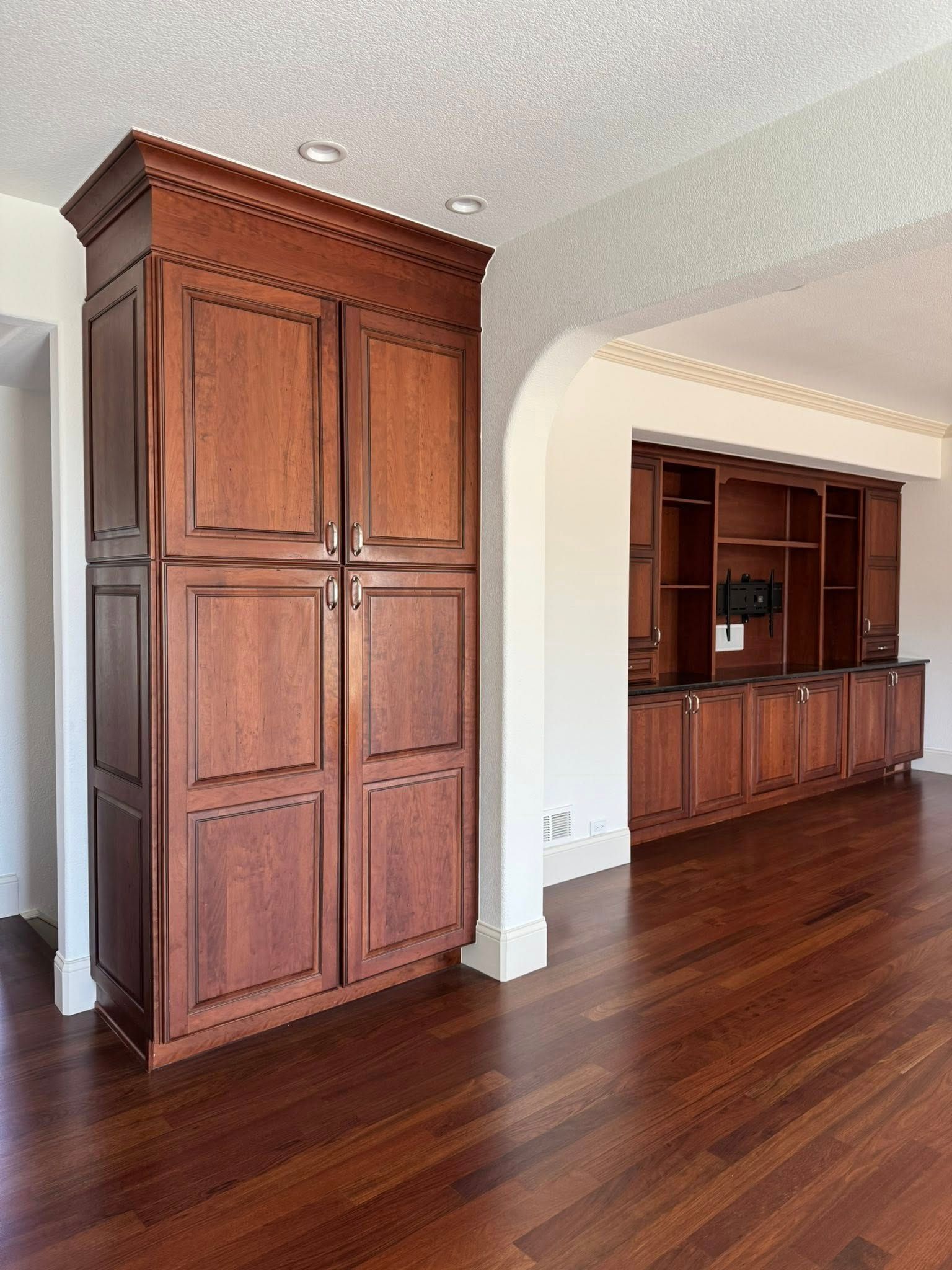 Tall, dark wood storage cabinet next to arched doorway and built-in wood shelves. Hardwood floor.