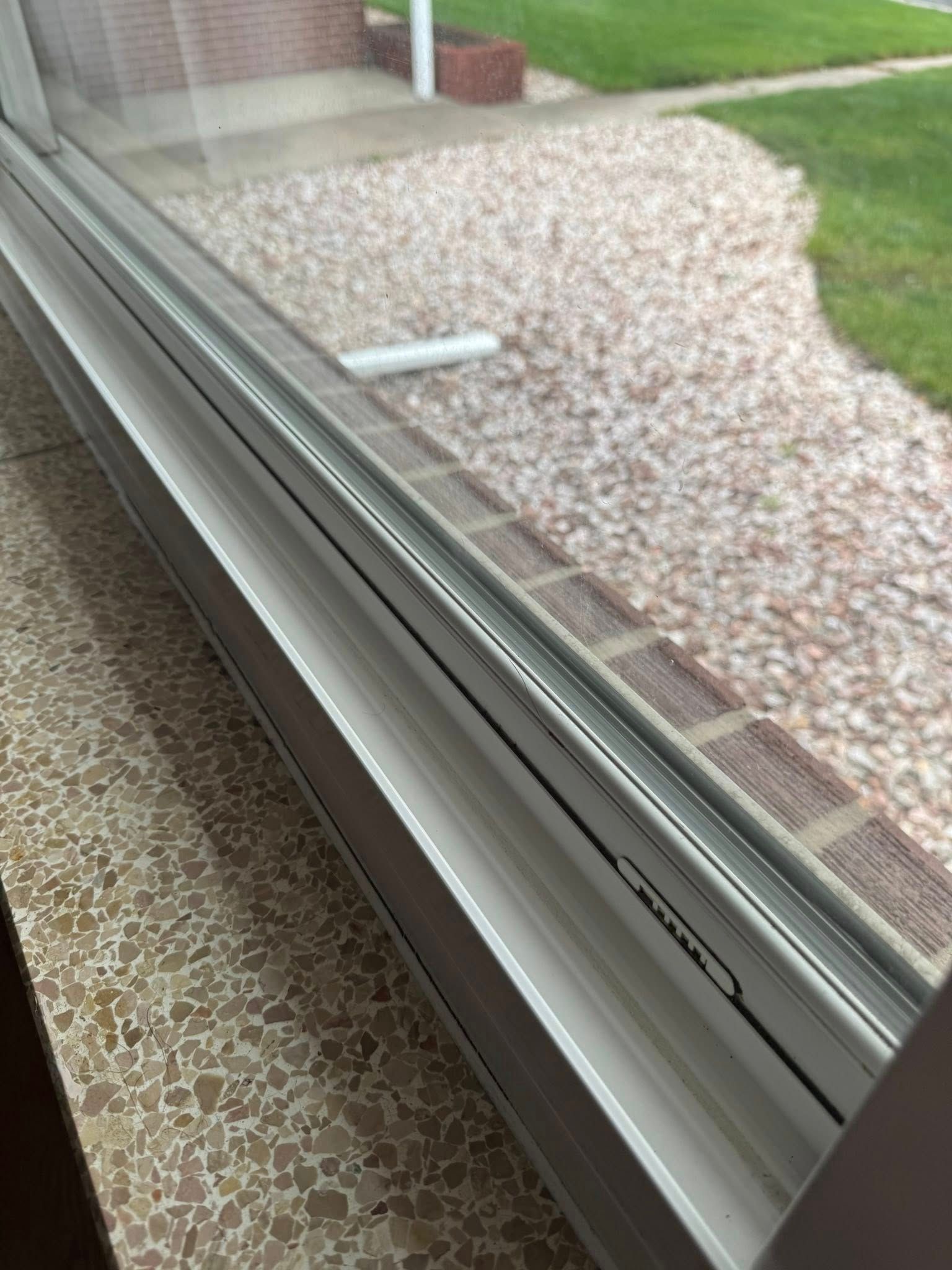 Window frame with a black line along the glass, looking out onto gravel, a lawn, and a brick building.