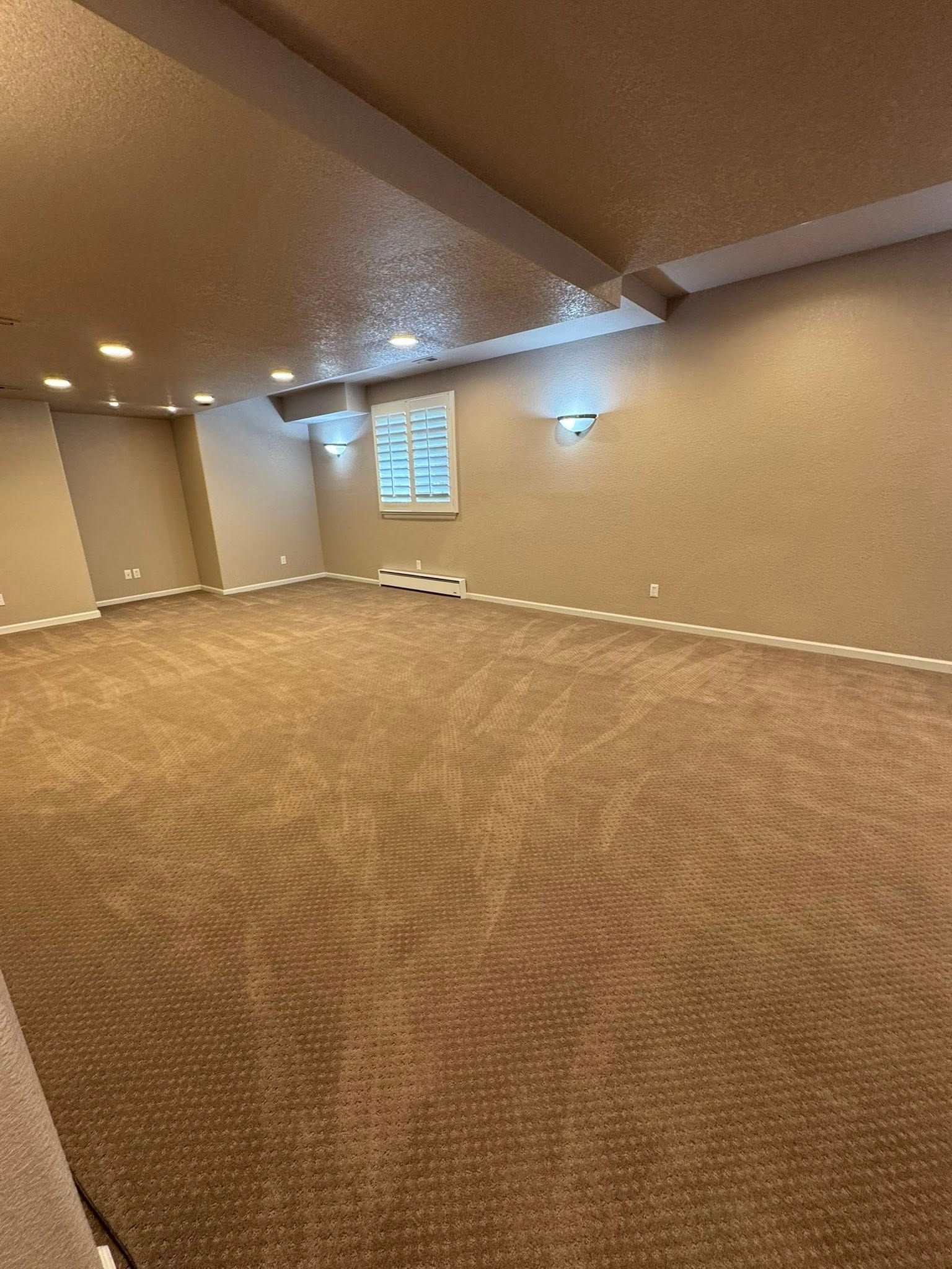 Empty room with brown carpet, tan walls, recessed lighting, and a small window with shutters.