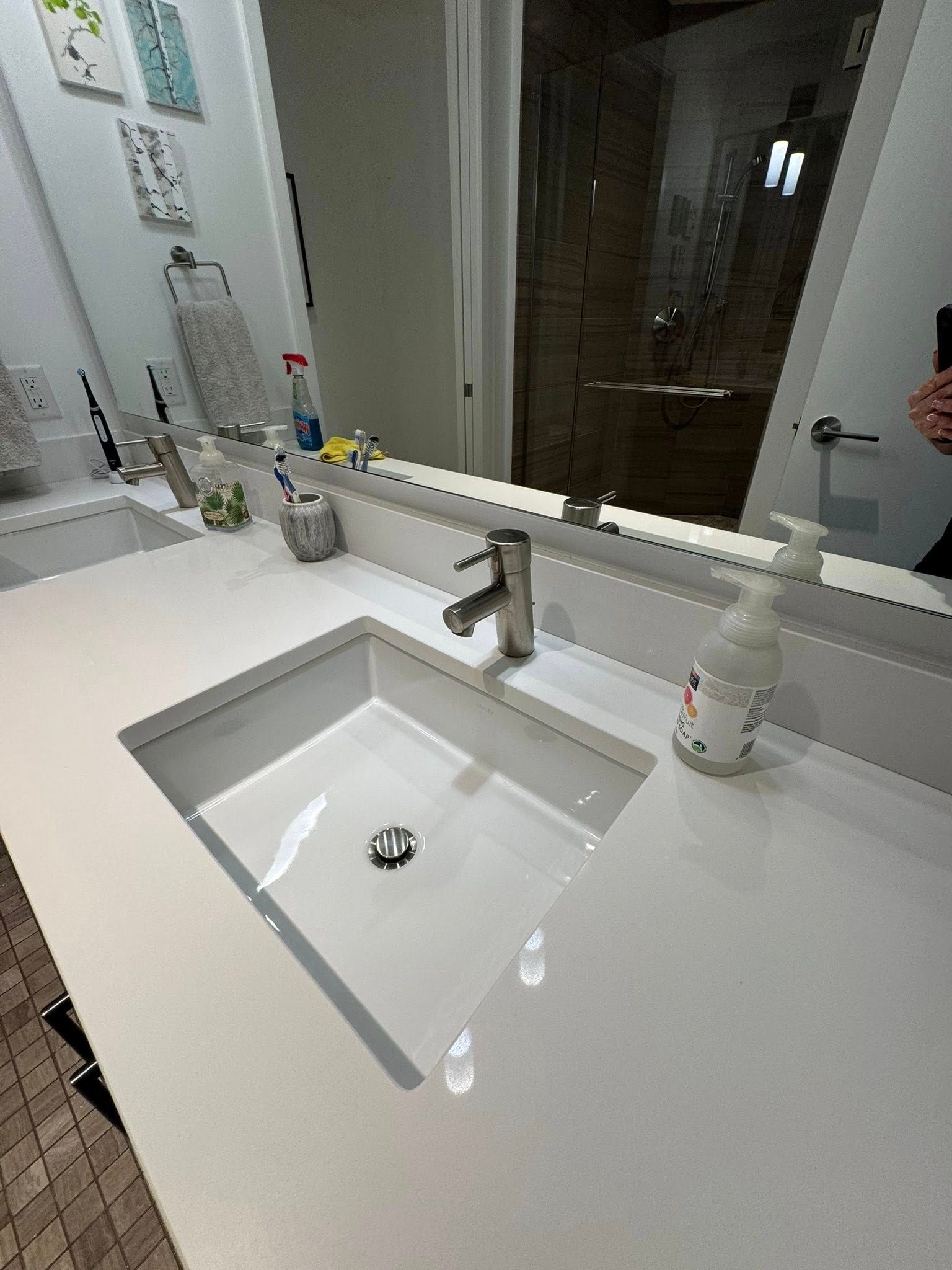 White bathroom sink with faucet and soap dispenser on a white countertop, with a mirror reflecting a shower.