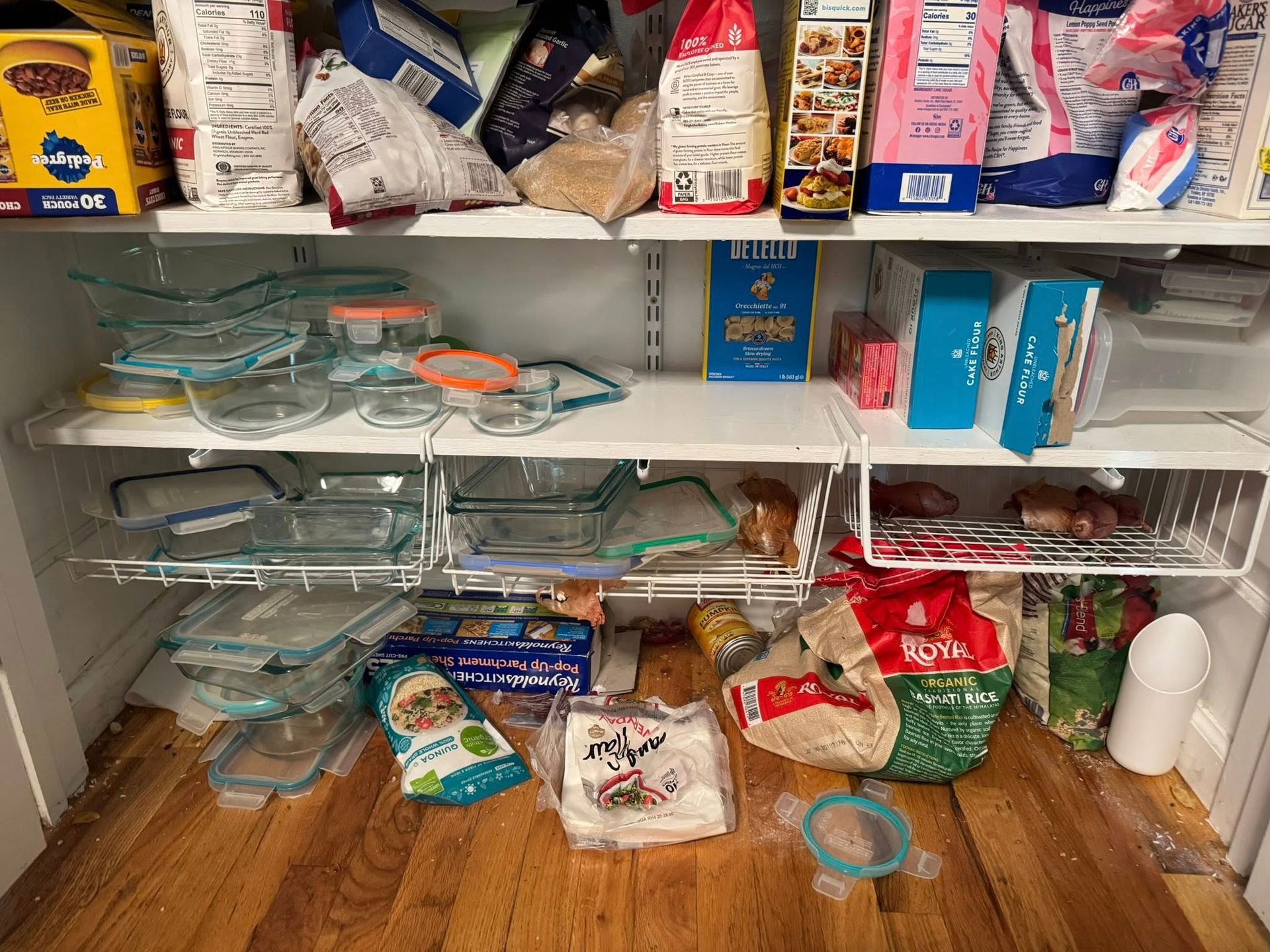A messy pantry with shelves holding various food items and glass dishes. Wooden floor.