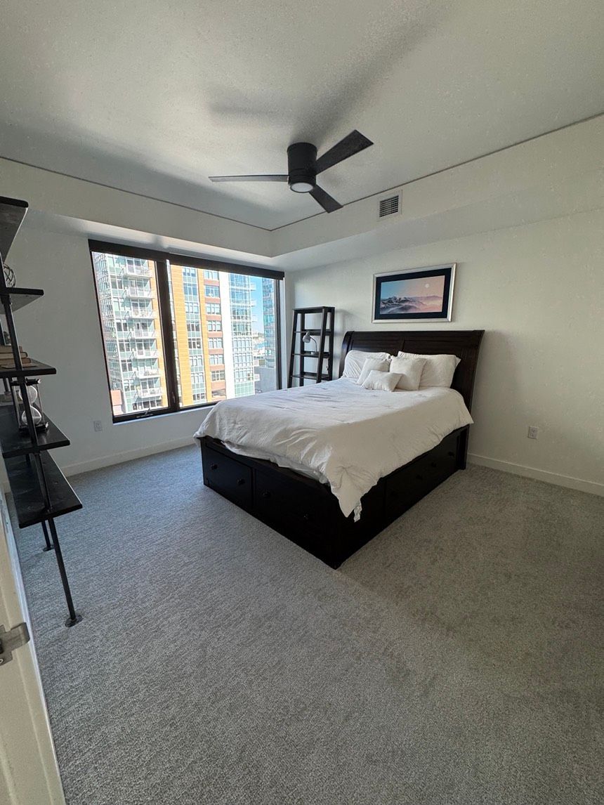 Bedroom with bed, shelves, and a window overlooking a cityscape. Gray carpet and white walls.