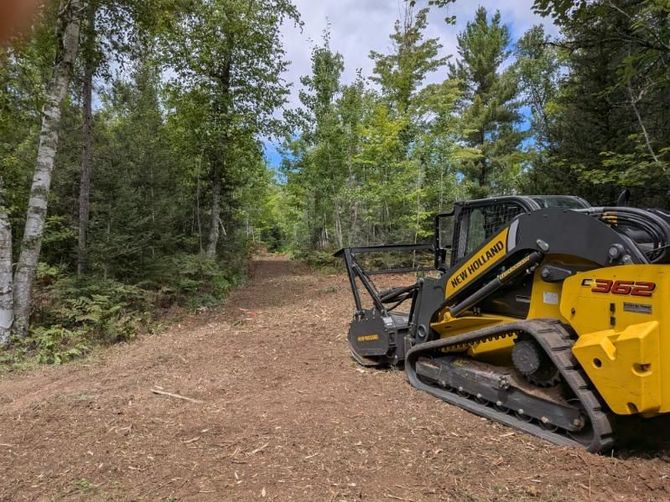 Yellow New Holland skid steer clearing a wooded path on a sunny day.