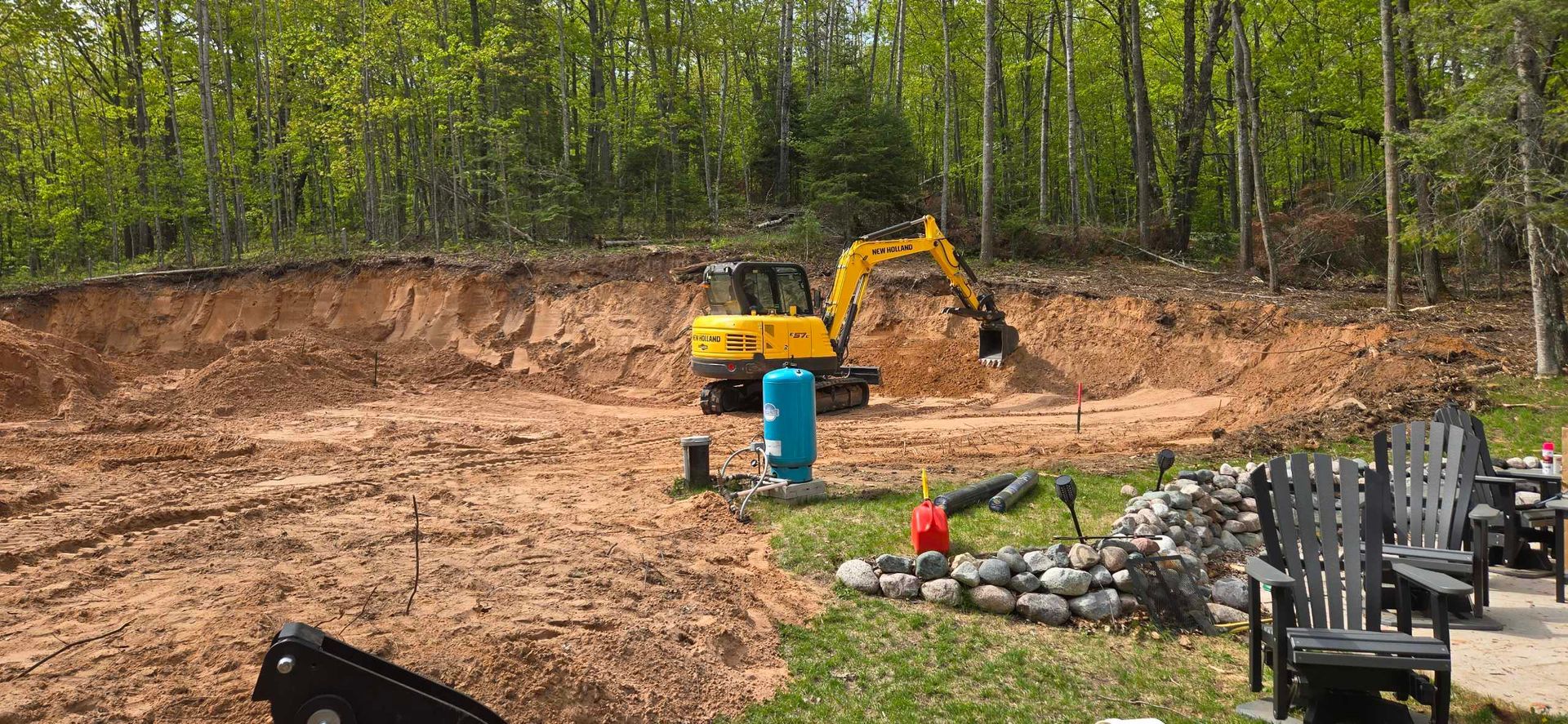 Yellow excavator digging a hole in a yard with forest background; Adirondack chairs visible.