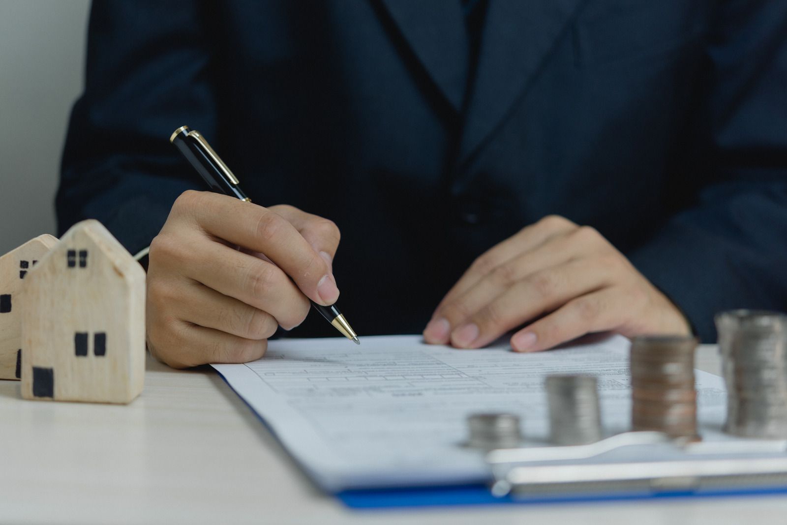 Person signing a document, miniature houses, and stacks of coins on a desk.