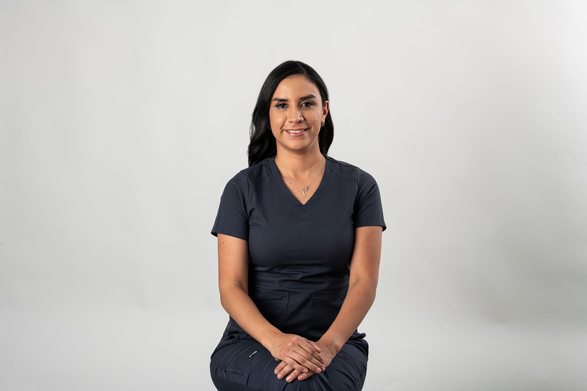 Woman in dark blue scrubs seated against a white backdrop, smiling, hands clasped.