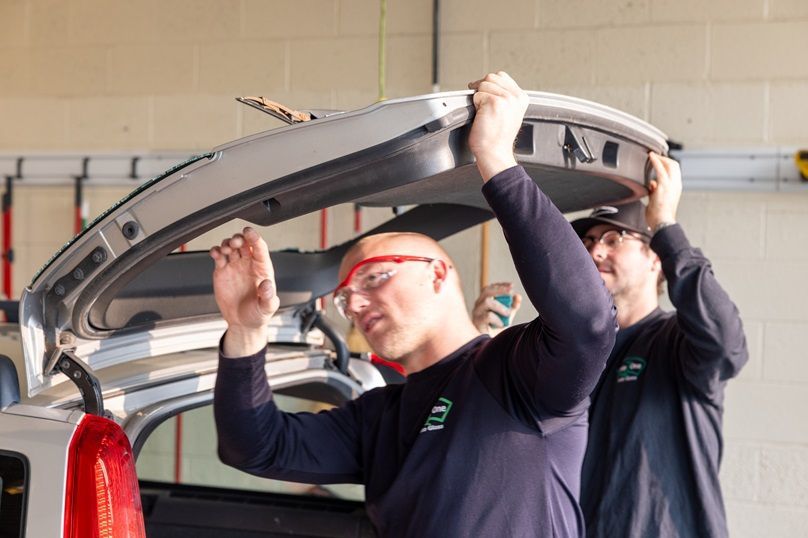 Two men are working on the trunk of a car in a garage.