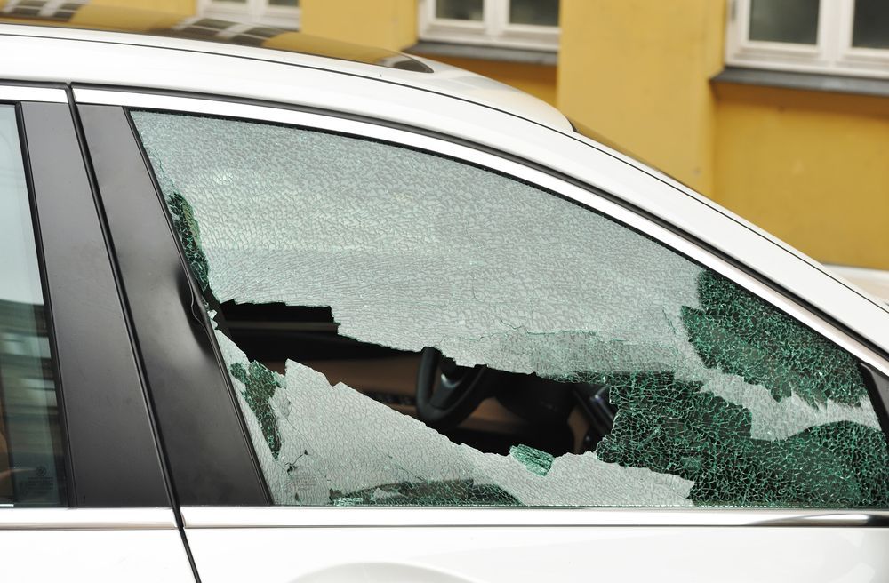 A car with a broken windshield is parked in front of a yellow building
