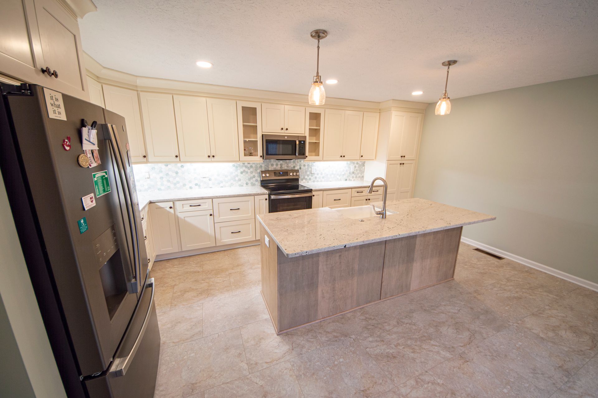 Modern kitchen with light cabinets, granite island, stainless steel appliances, and beige flooring.