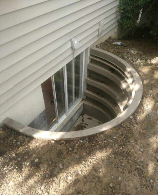 Basement window with a concrete well. Beige siding, dirt, and a concrete semi-circular stairwell.