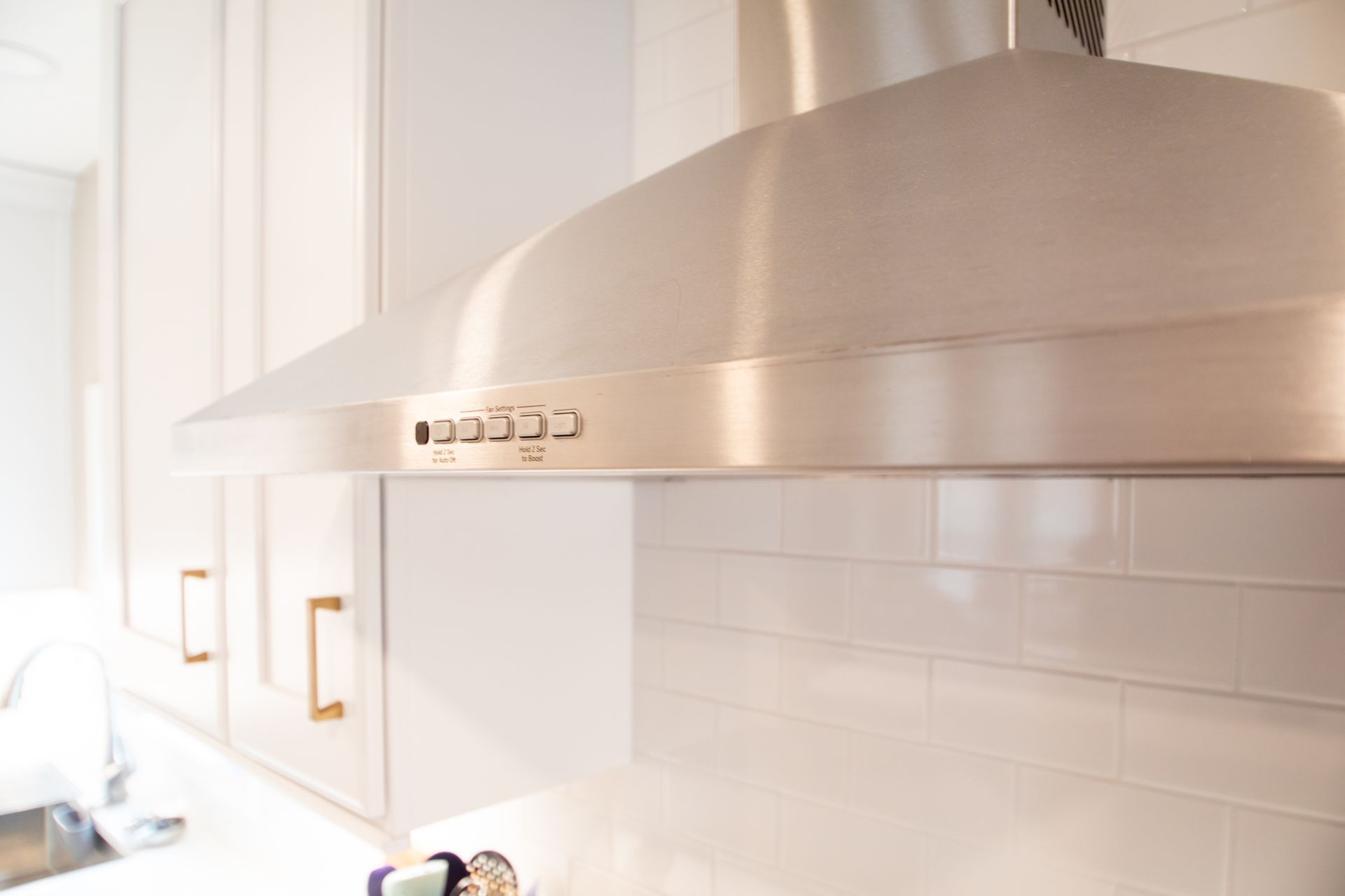 Stainless steel range hood over a white tiled backsplash and cabinets with gold handles.