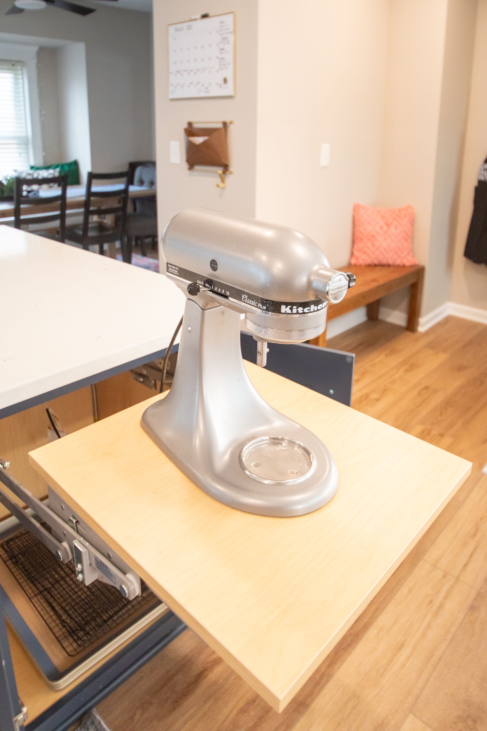 Silver stand mixer on a pull-out wooden shelf in a kitchen; island, dining table and bench in background.