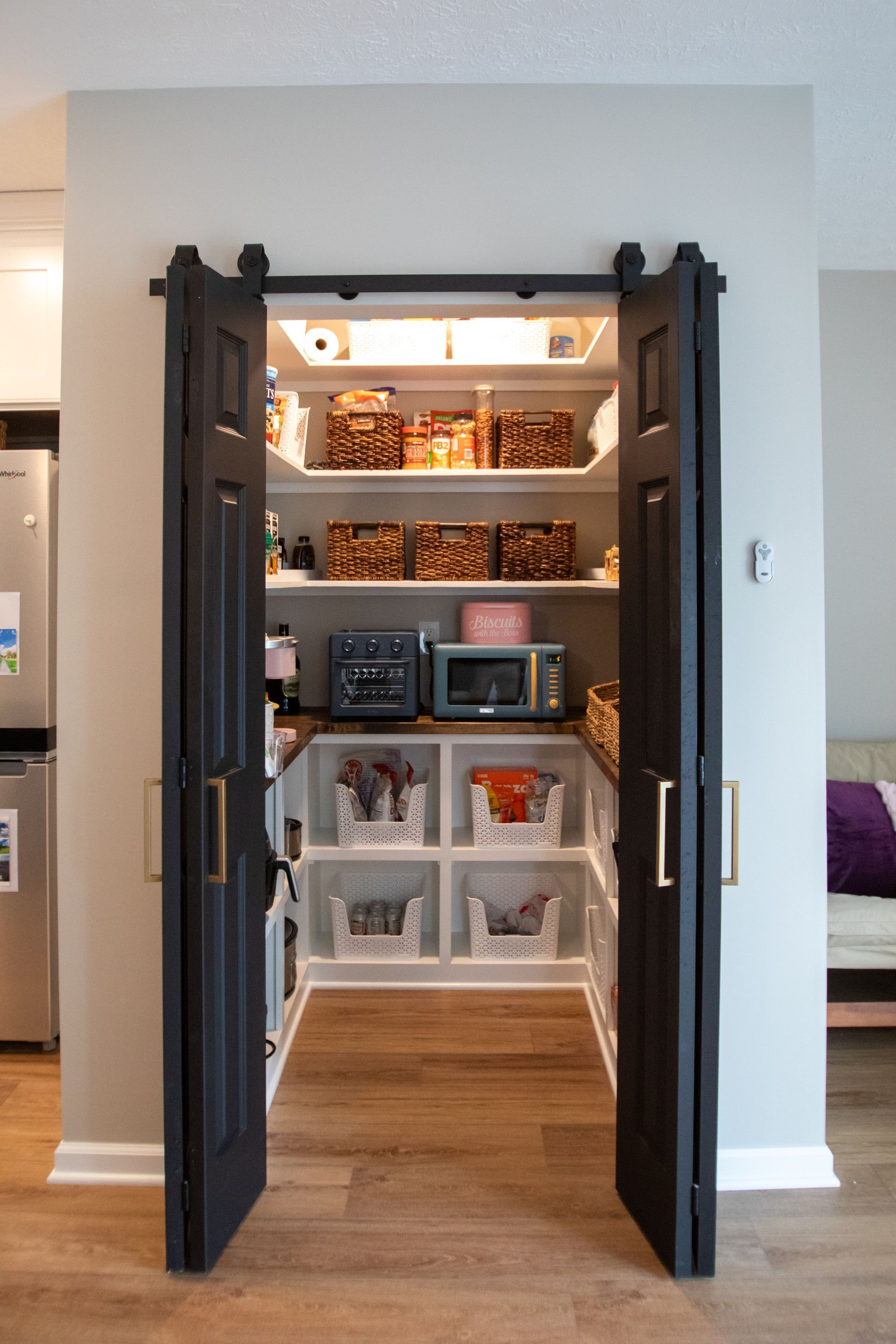 Black barn-style doors open to a well-organized pantry with various food items, small appliances, and storage bins.