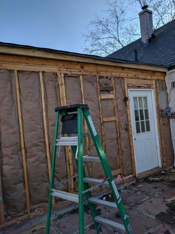 Construction site with exposed wooden frame, insulation, and a ladder next to a door.